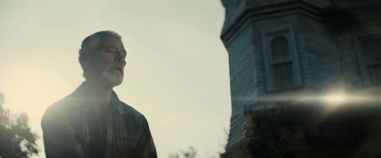 Don’t Breathe 2 (2021) movie still: medium — A man stands outdoors near the weathered wooden exterior of a tall suburban house. The low sun creates…