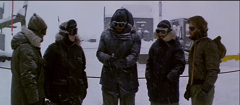 The Thing (1982) movie still: fullbody — An arctic research base exterior during a snowstorm features white snow and metallic structures. Five men…