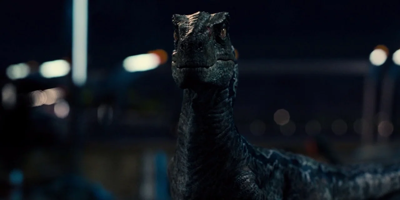 Jurassic World (2015) movie still: closeup — A dark outdoor lot at night with blurred industrial lights in the background. A velociraptor stands in…