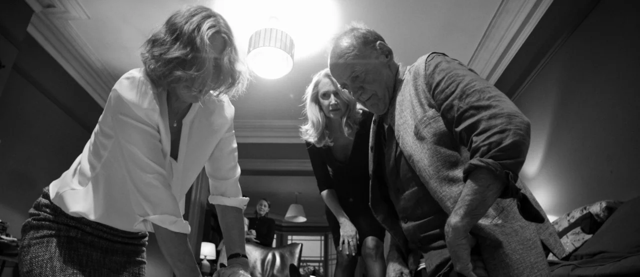 The Party (2017) movie still: medium — A dimly lit room with a central overhead light casting strong shadows on the subjects. Three adults stand in…