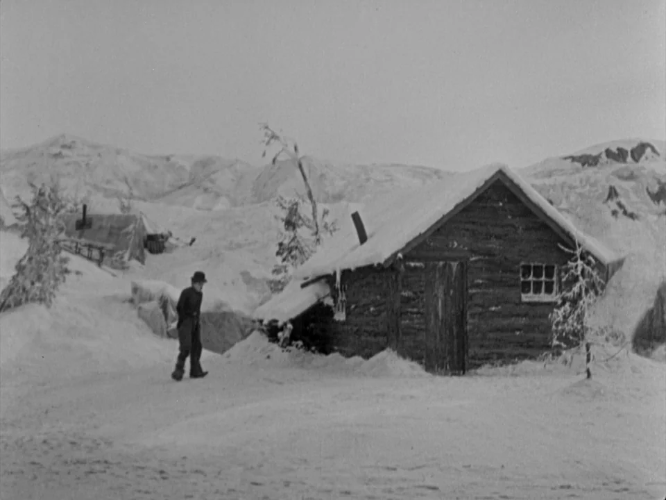 The Gold Rush (1925) movie still: wide — A snowy, barren mountain landscape features a small log cabin partially buried in snow. A small man in a…