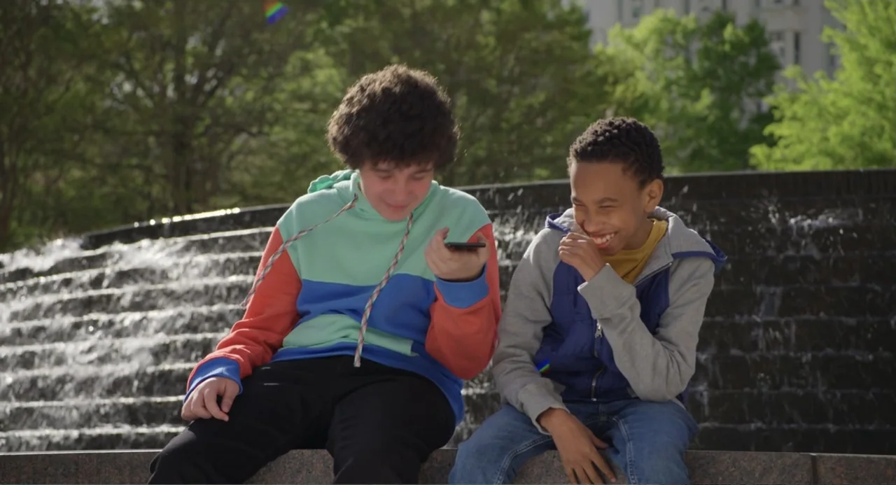 Harold and the Purple Crayon (2024) movie still: medium — Two teenage boys sit on the stone ledge of a public tiered water fountain. The boys are…
