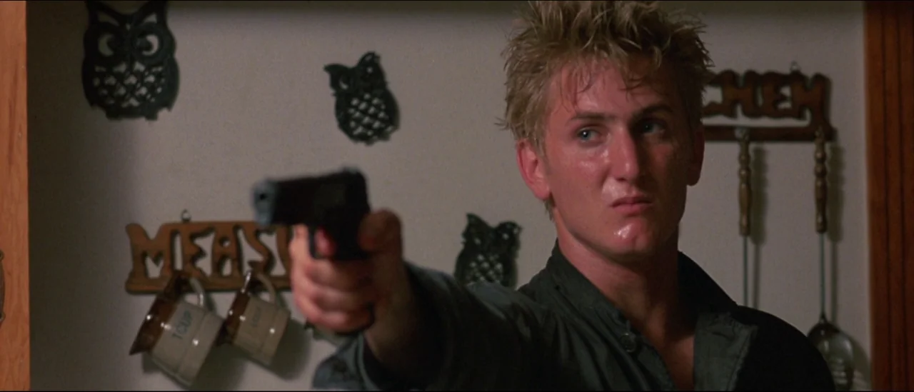 At Close Range (1986) movie still: medium — A wall decorated with wooden cutouts of owls and measuring cups in a room. A young man holds a black…