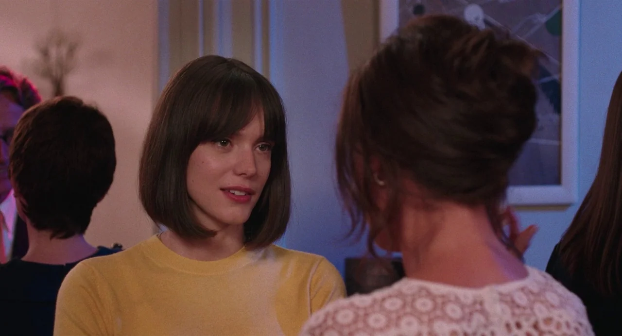 Godard Mon Amour (2015) movie still: medium — A group of people gather in a softly lit interior space during an evening social. A young woman with a…