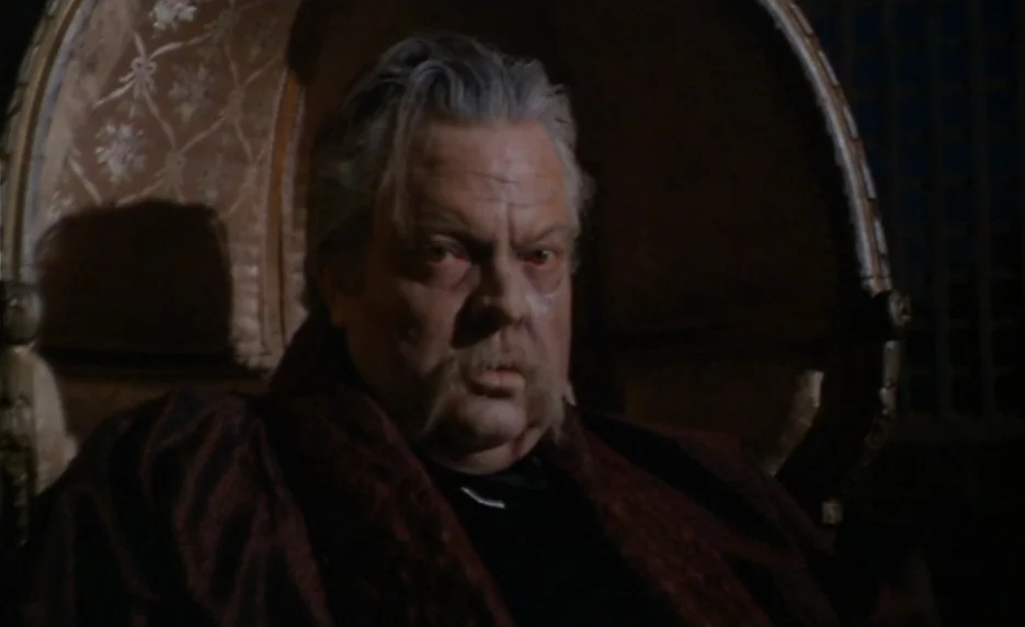 The Immortal Story (1968) movie still: closeup — An older man sits in a large, upholstered high-backed chair in a dimly lit room. The wall behind…