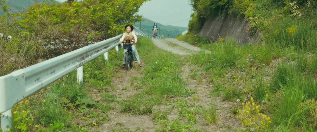 Monster (2023) movie still: wide — A rural dirt path lined with tall grass and a silver metal guardrail. Two young people ride bicycles toward the…