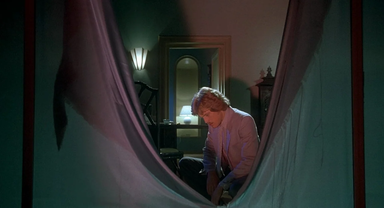 Inferno (1980) movie still: medium — A dimly lit room with draped fabric in the foreground partially obscuring the view. A man kneels on the floor…