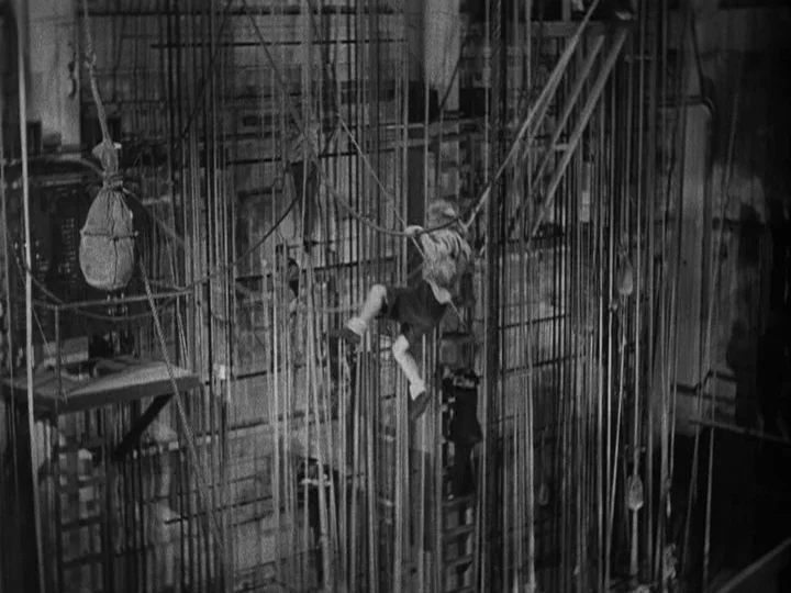 A Night at the Opera (1935) movie still: wide — A complex web of ropes and wooden battens fills the dark theater fly loft. A young boy holds onto a…