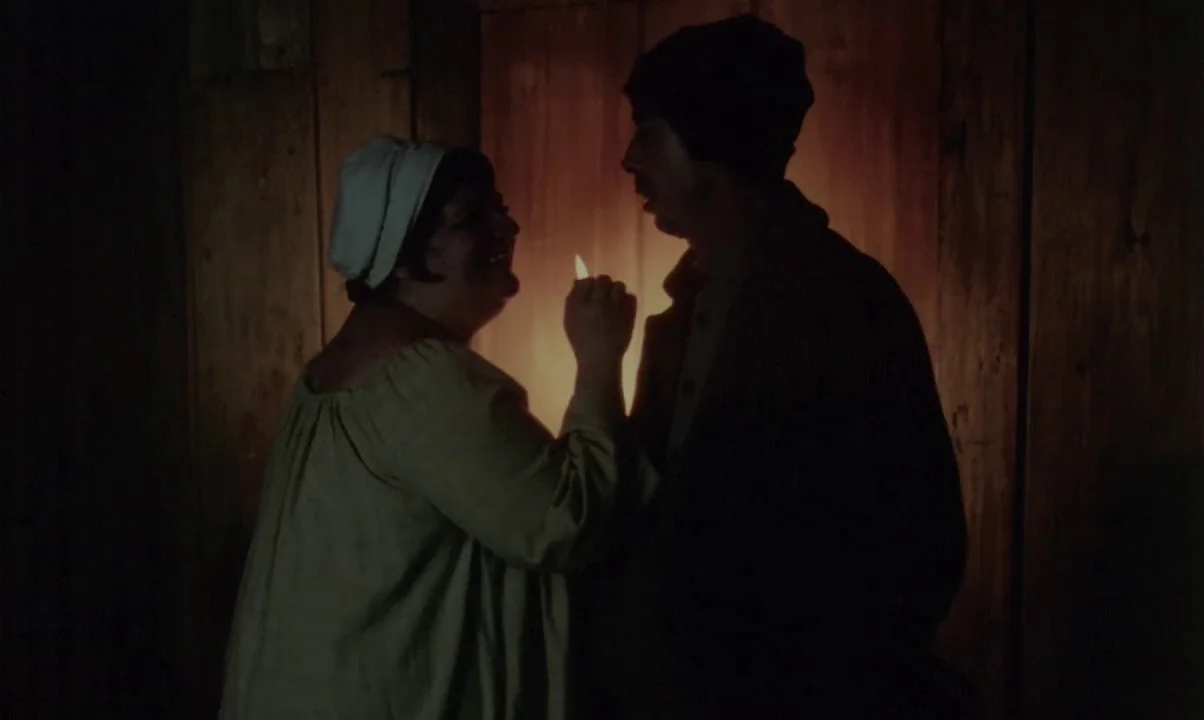 Kamouraska (1973) movie still: medium — A dark, cramped room featuring vertical wooden planks on the back wall. Two individuals stand close together…