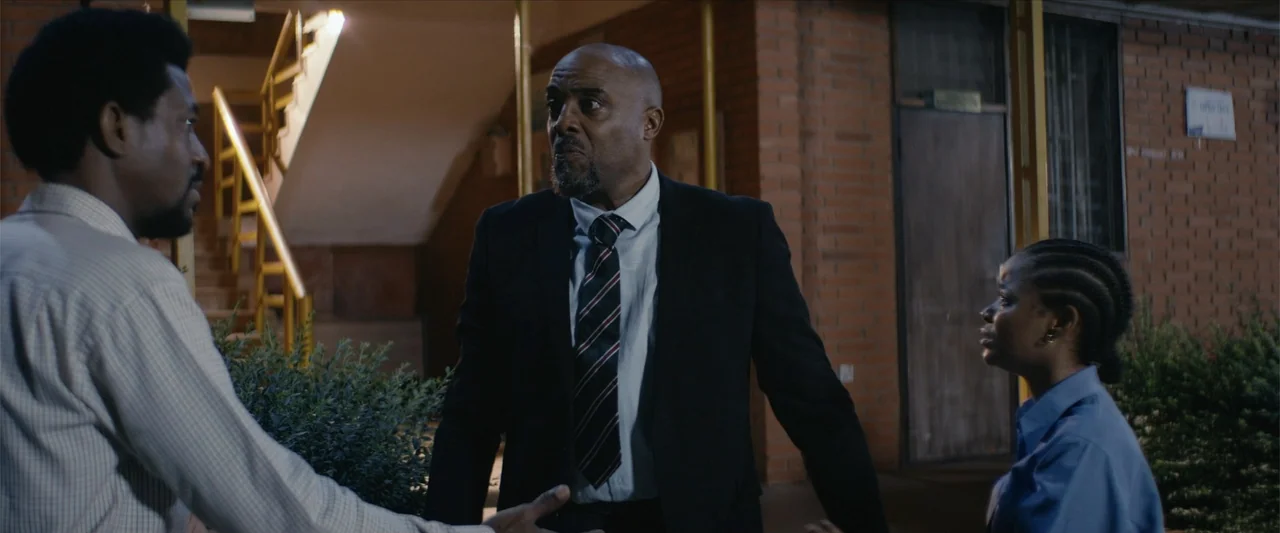 Ms. Kanyin (2025) movie still: medium — An outdoor passageway at night features exposed brick walls and a metal staircase railing. Three individuals…