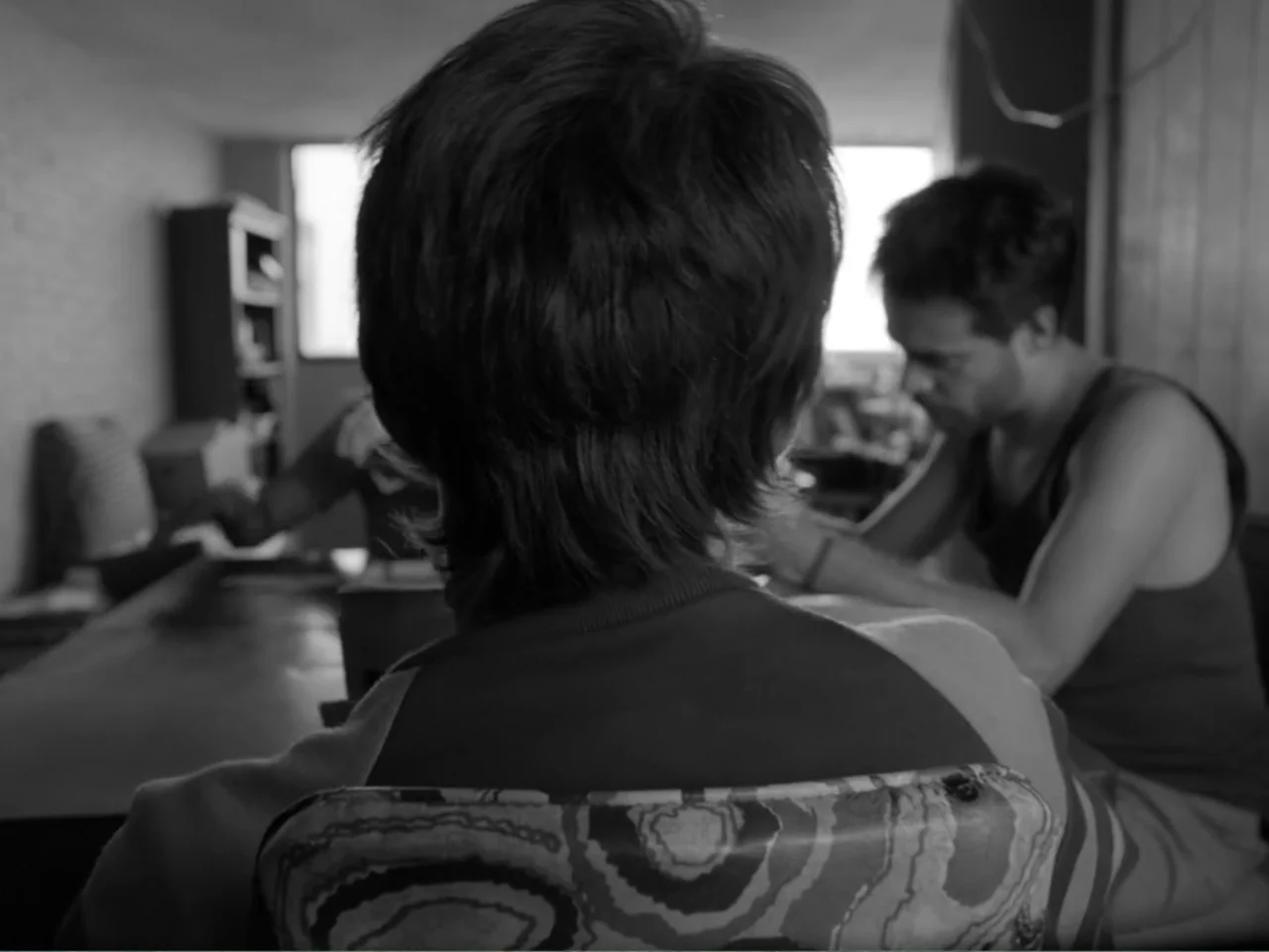 Güeros (2014) movie still: medium — A dimly lit room with a desk and chairs placed near a bright window. Two young men are present while one is…