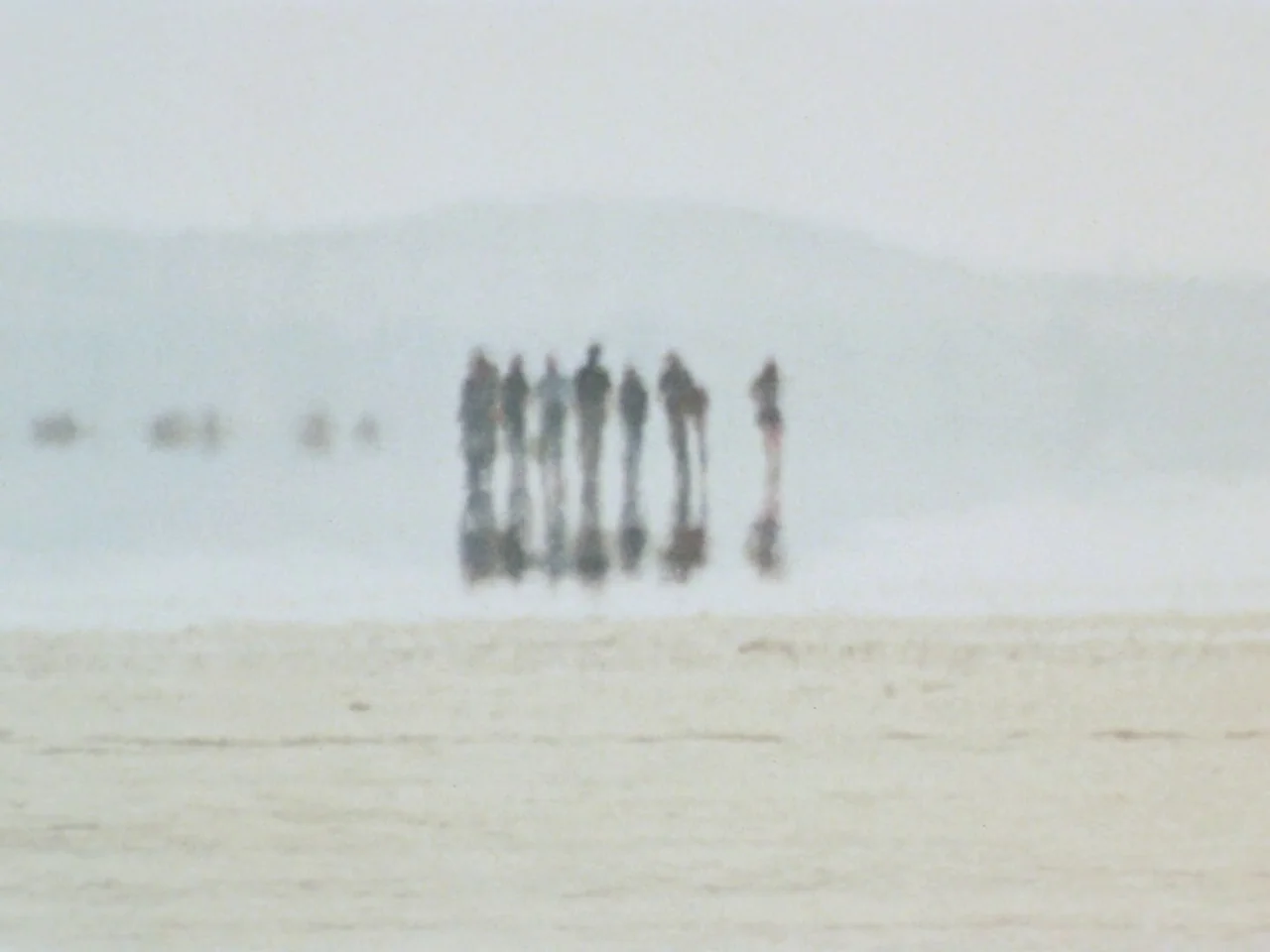 Punishment Park (1971) movie still: wide — A vast, light-colored sandy beach stretches out under a hazy, washed-out sky. A small, blurry group of…