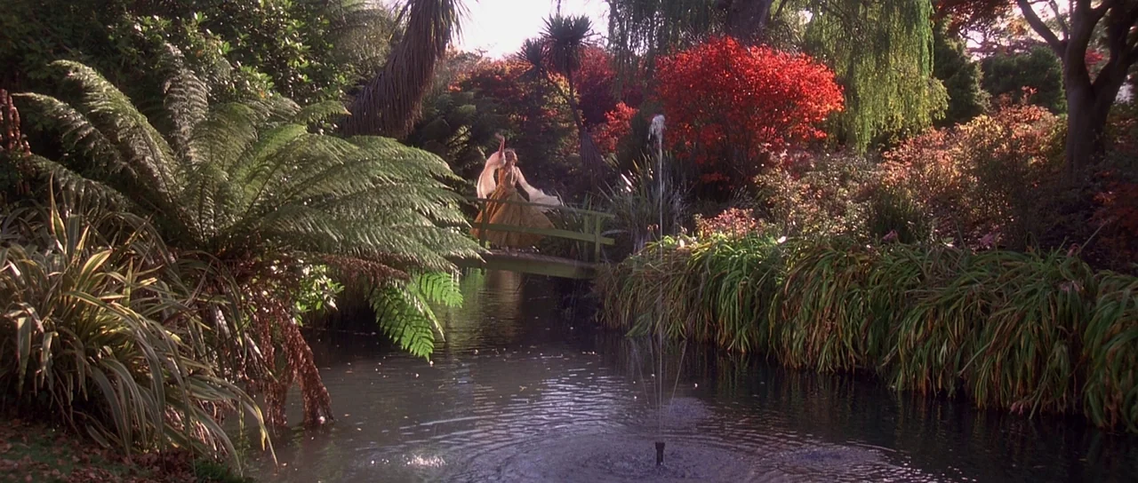 Heavenly Creatures (1994) movie still: wide — A lush botanical garden surrounds a tranquil stream with a small wooden footbridge. A person in a long…