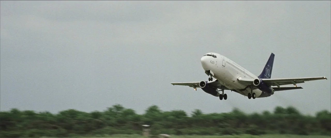 Blood Diamond (2006) movie still: wide — A commercial jet aircraft ascends from a concrete runway against a grey overcast sky. Green trees line the…