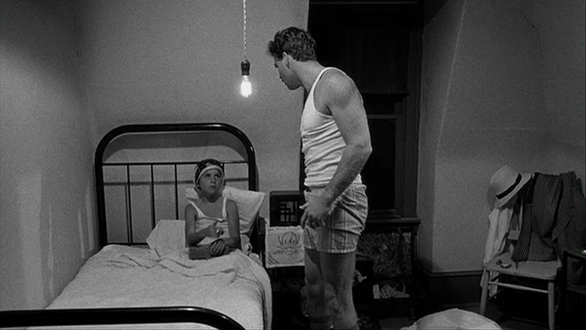 Paper Moon (1971) movie still: medium — A small attic with sloped walls and a single bare lightbulb illuminating the space. A man stands facing a…