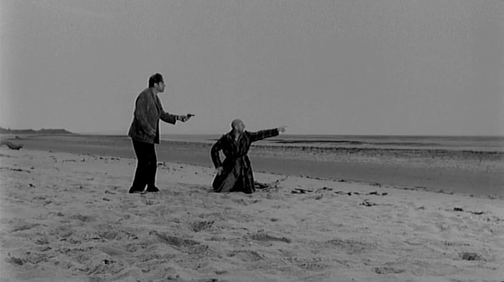 Cul-de-sac (1966) movie still: wide — A wide, flat expanse of sandy beach stretches under a pale, overcast sky. One man kneels and points seaward as…