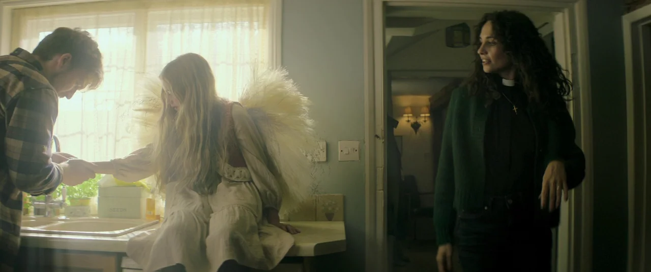 Lord of Misrule (2023) movie still: medium — A sunlit kitchen has a young girl with large white feathery wings sitting on counter. A man reaches…