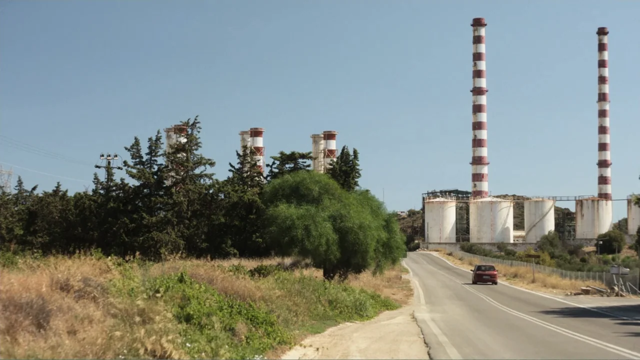 Hot Milk (2025) movie still: wide — A rural road winds past tall red-and-white striped industrial smokestacks and storage tanks. A small red car…