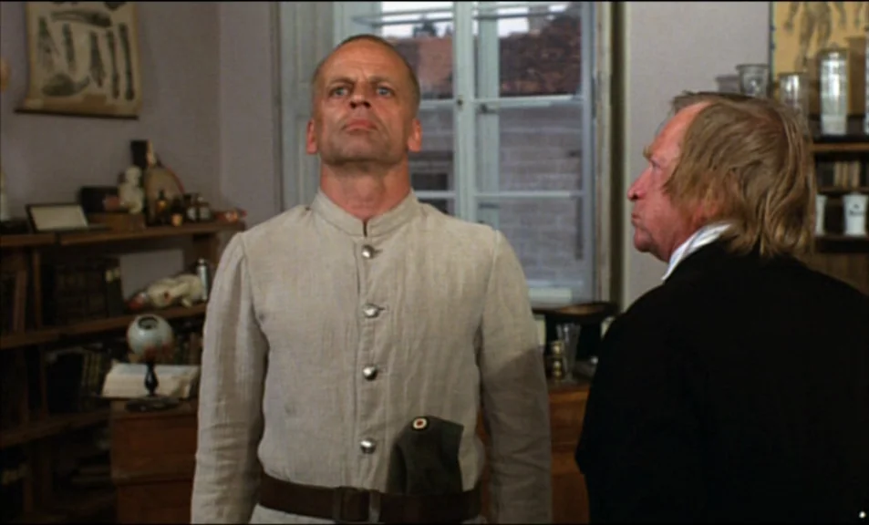 Woyzeck (1979) movie still: medium — A medical or academic study features wooden shelves filled with glass jars and books. A bald man wearing a…