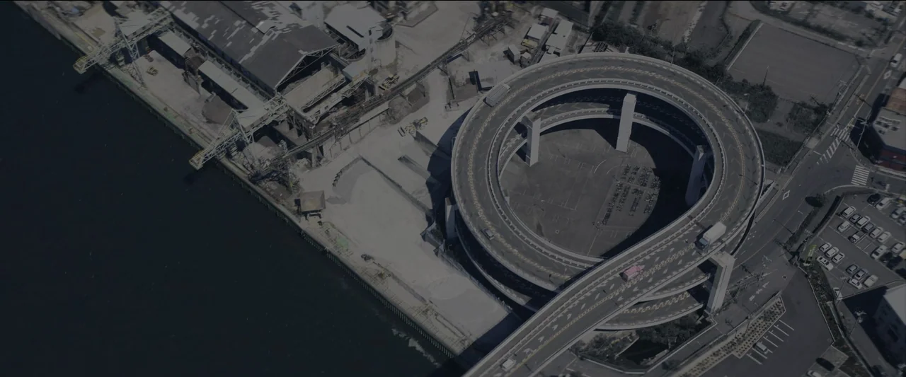 Kate (2021) movie still: establishing — A high-angle view shows a complex circular elevated highway ramp next to industrial docks. A large grey…