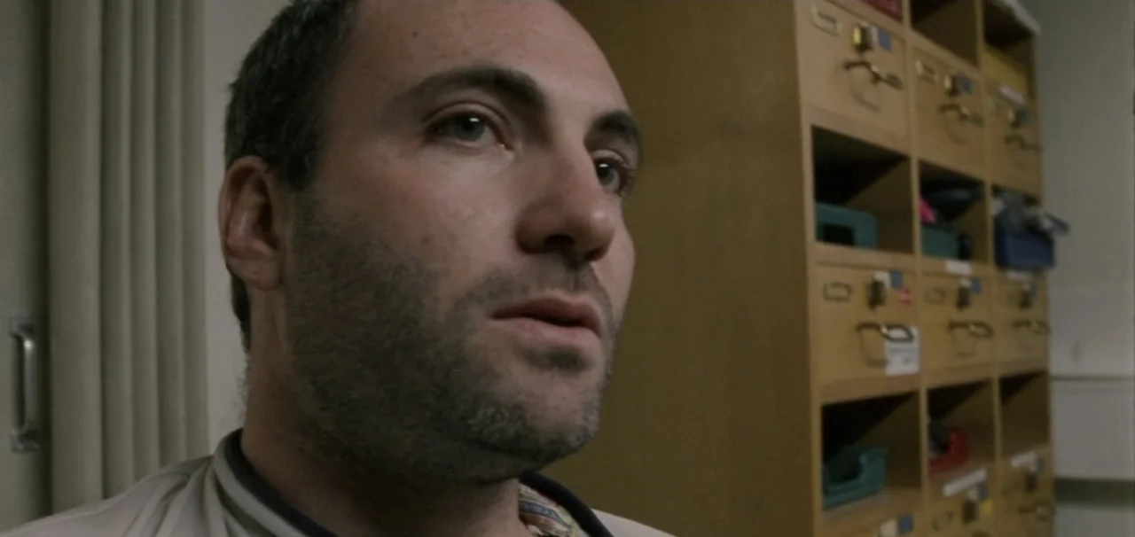 Pusher (1996) movie still: closeup — A utilitarian office space with wooden grid shelving containing various plastic storage bins. A man in a light…