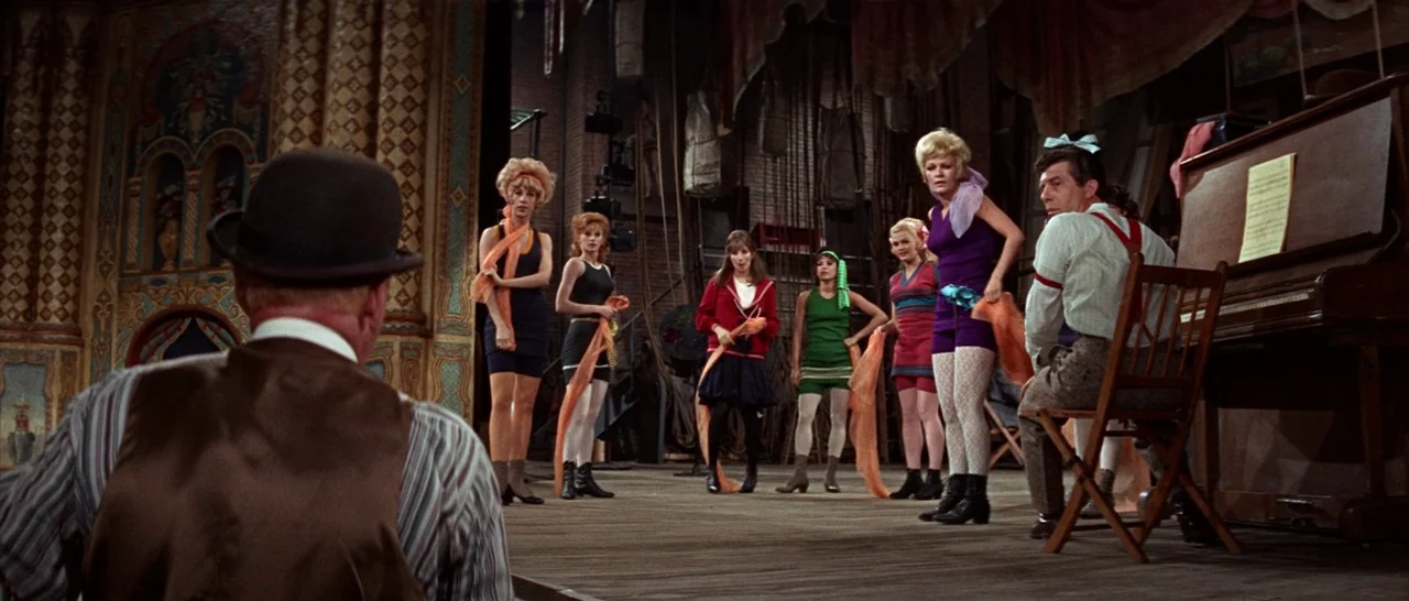 Funny Girl (1968) movie still: wide — A wooden stage set with ornate wall panels and a piano sits inside a theater. A group of women in 1920s-style…