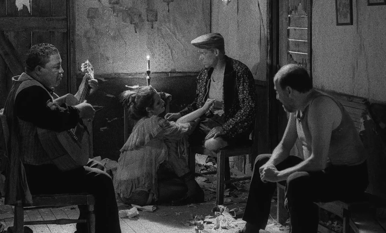 Go Down Death movie still: wide — A dimly lit room with peeling walls and wooden floorboards contains scattered debris. Four people sit together in…
