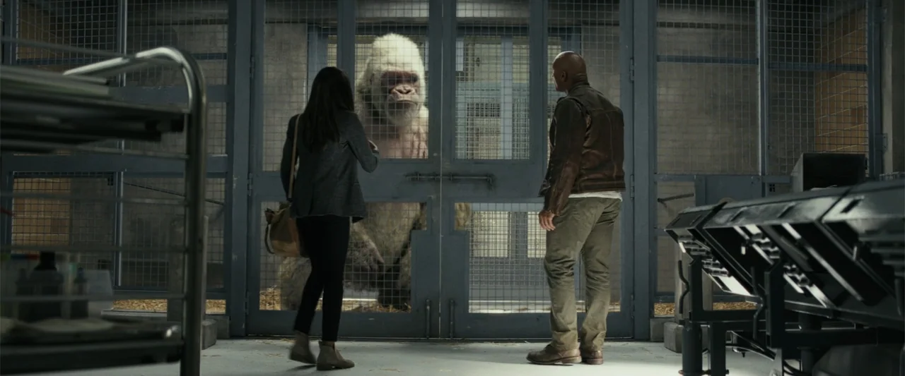 Rampage (2018) movie still: fullbody — A sterile industrial facility contains a large metal cage with hay on the floor. A man and a woman face the…