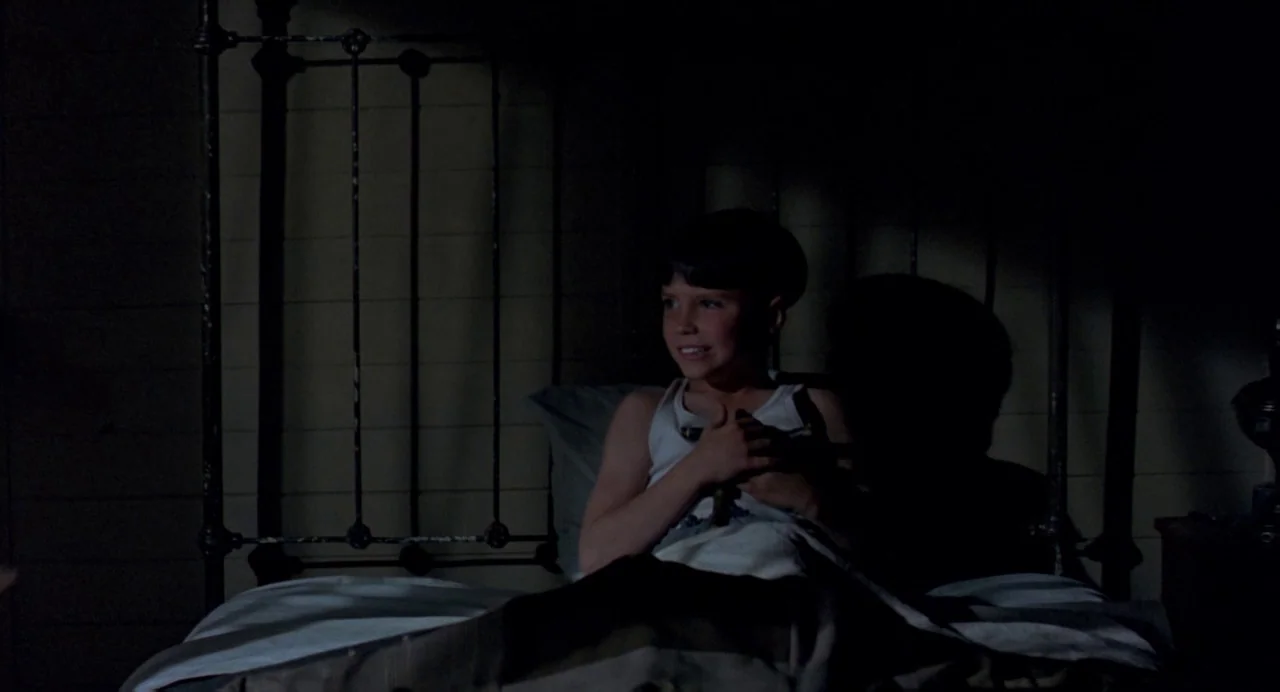 The Reflecting Skin (1990) movie still: medium — A child sits up in an iron-framed bed inside a dark room at night. The boy wears a white tank top…