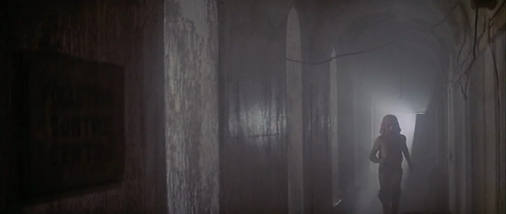 Zardoz (1974) movie still: wide — A long concrete corridor is filled with dense smoke and arched alcoves along walls. A man runs towards the camera…