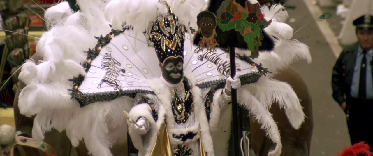 JFK (1991) movie still: medium — A man in an elaborate Mardi Gras Zulu parade costume stands outdoors. He holds a staff topped with a decorated…