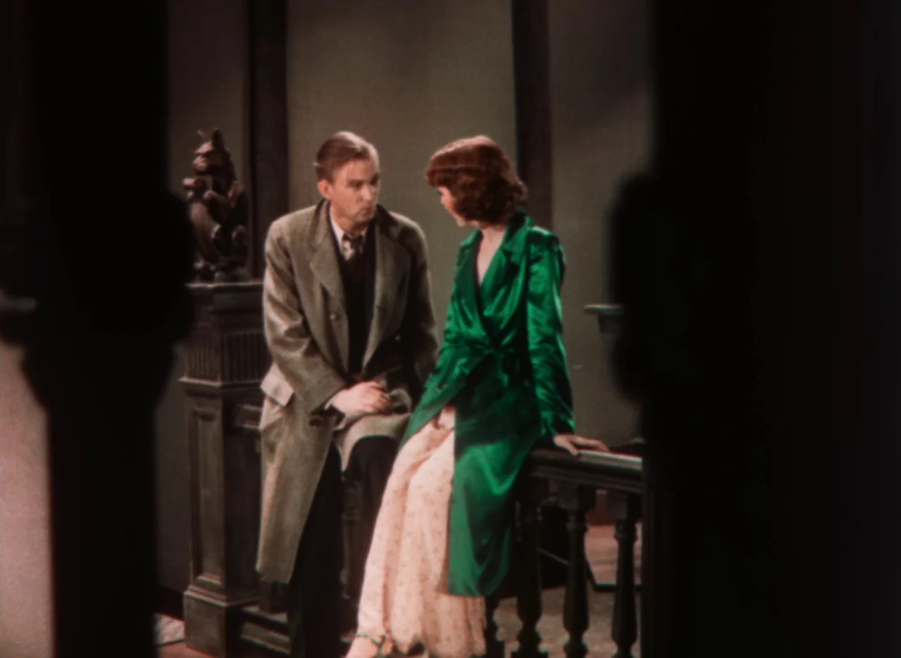 Doctor X (1932) movie still: medium — A man and woman sit on the wooden banister of a dark staircase interior. The man wears a tweed coat and the…