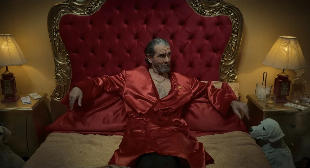 The Beauty and the Moody (2022) movie still: medium — A lavish bedroom features a massive red upholstered headboard with gold-trimmed edges. A…