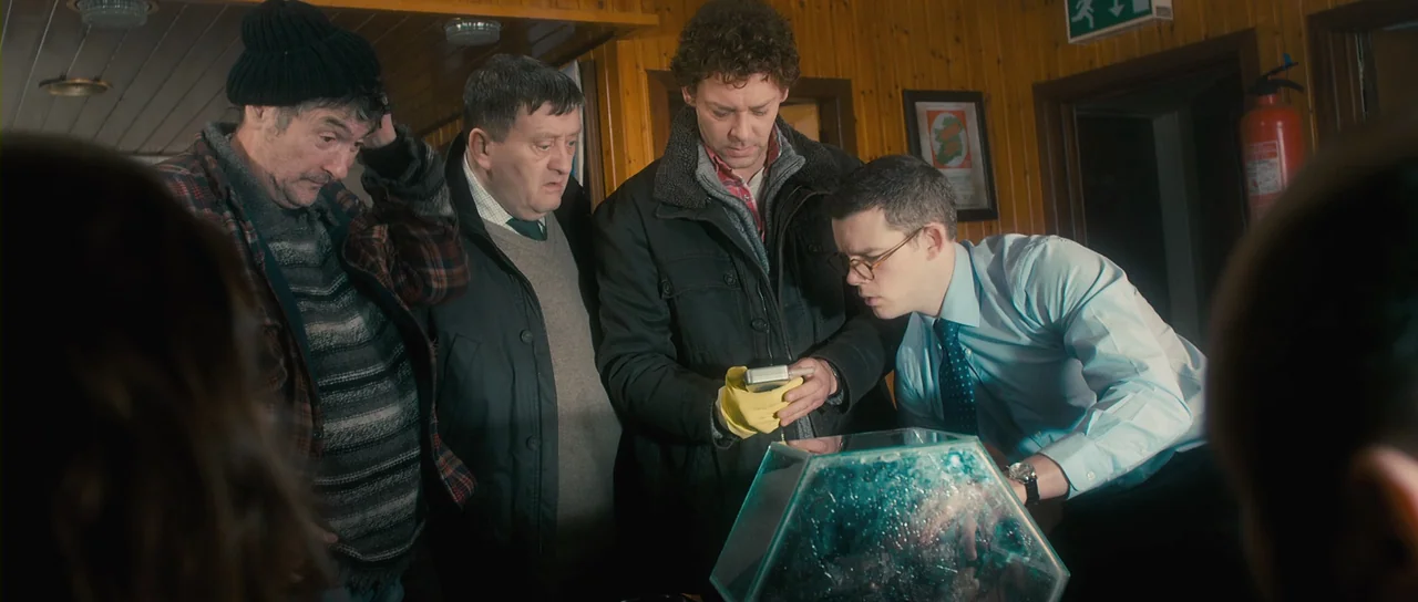 Grabbers (2012) movie still: medium — A wood-paneled room contains four men gathered around a low glass display case. The men lean forward in a…