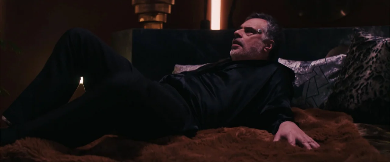 M3GAN 2.0 (2025) movie still: medium — A dimly lit room with a brown faux-fur bedspread and dark walls. A middle-aged man with a temple implant lies…