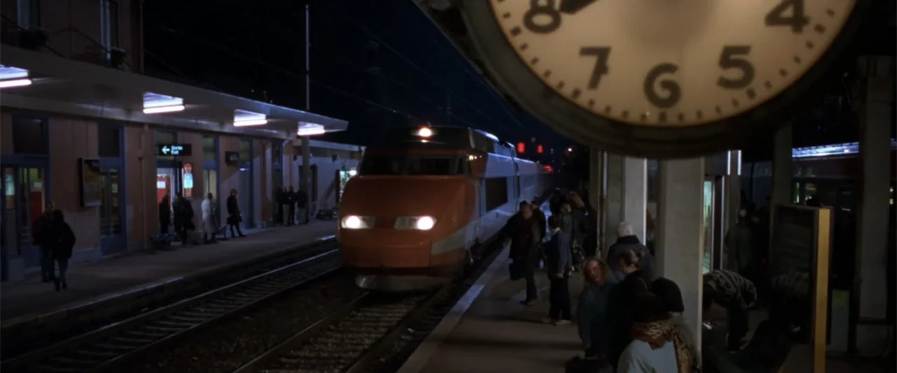 Maximum Risk (1996) movie still: wide — A night view of a train station platform with a modern passenger train approaching. Commuters stand along…