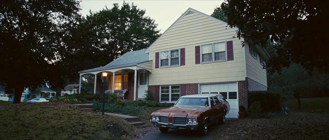 The Lovely Bones (2009) movie still: establishing — A two-story suburban house features yellow siding and a brick lower level at dusk. An orange…