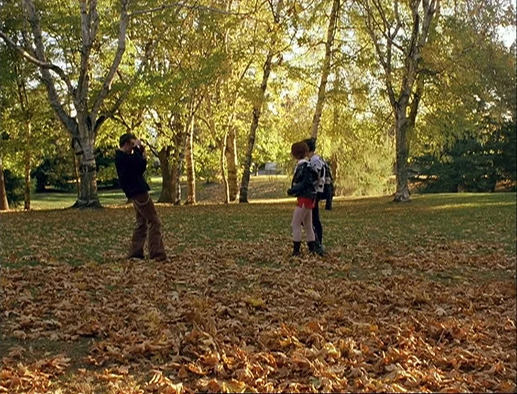 Elephant (2003) movie still: wide — A leaf-covered public park area features tall deciduous trees with yellow autumn foliage. A man stands holding a…