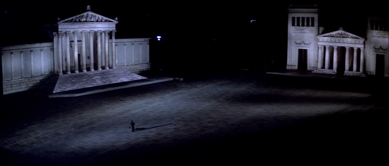 Suspiria (1977) movie still: establishing — A vast, paved plaza at night is flanked by two large neoclassical stone buildings. A single person…