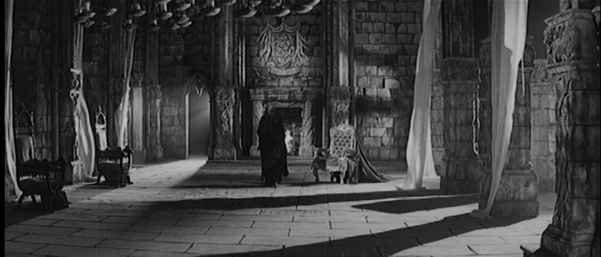 I Vampiri (1957) movie still: wide — A stone chamber features massive columns, thin fabric drapes, and a central fireplace. An elderly figure in a…