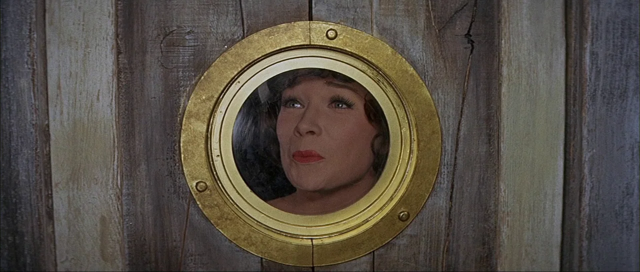 What A Way To Go! (1964) movie still: closeup — A woman looks through a circular brass porthole mounted in a wooden wall panel. The porthole acts as…