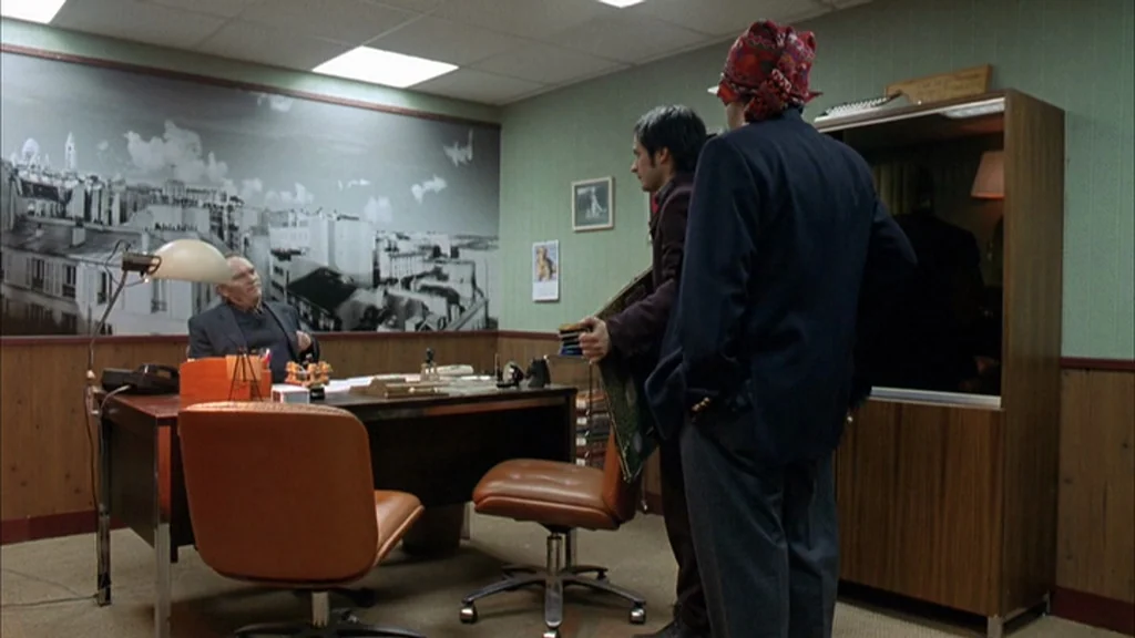The Science Of Sleep (2006) movie still: wide — An office with light green walls features a large photographic mural of a city. An older man sits at…