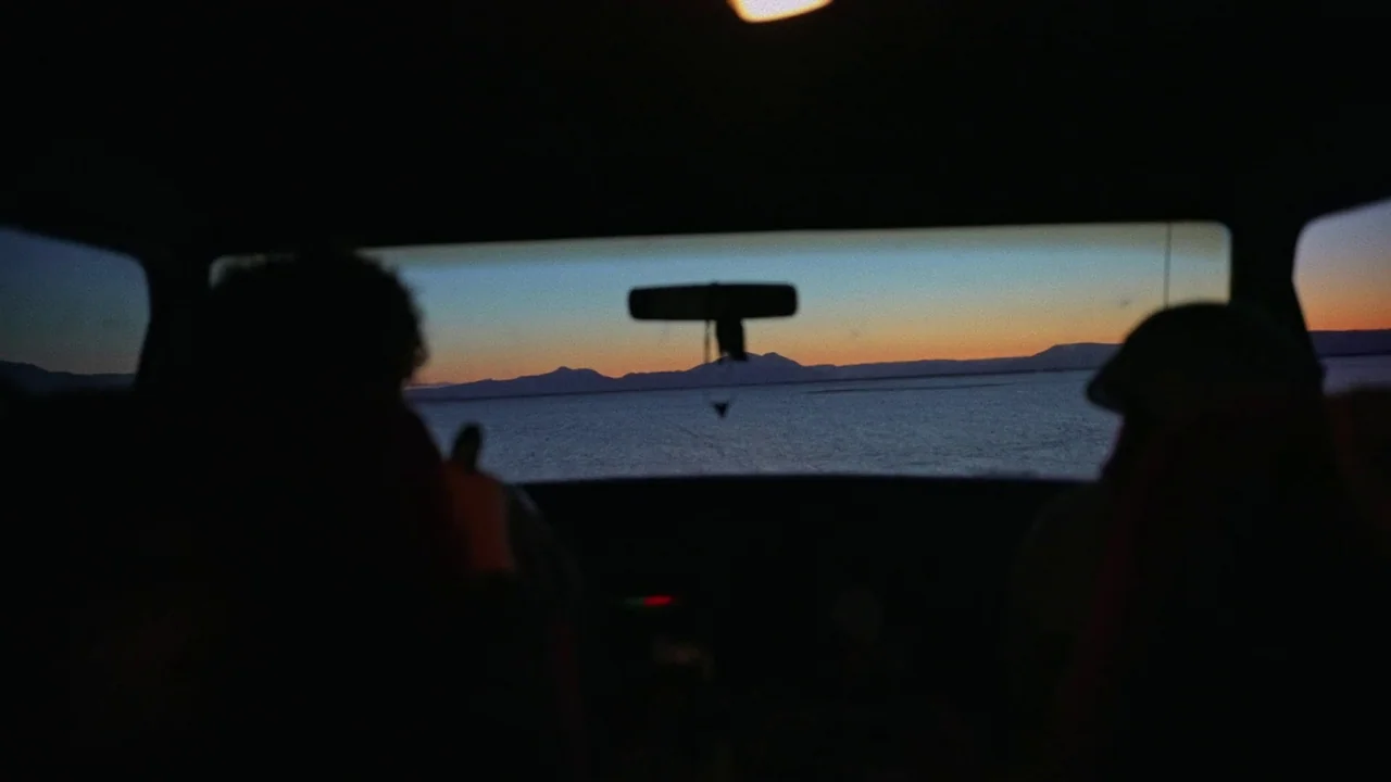 Gasoline Rainbow (2023) movie still: medium — The interior of a dark car faces out towards a distant horizon at twilight. Two silhouetted figures…