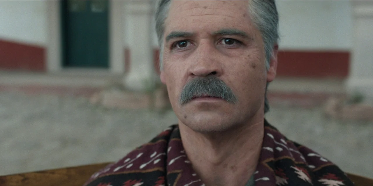 Pedro Páramo (2024) movie still: closeup — An outdoor area stands before a building with a white and maroon wall facade. A middle-aged man with a…