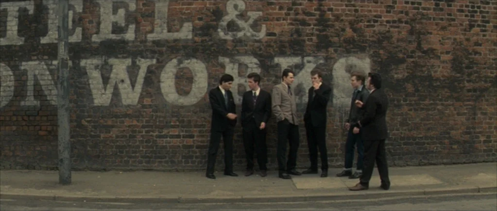 Nowhere Boy movie still: wide — A group of six men in suits stand in front of a brick wall. The wall is weathered with faded white paint that spells…