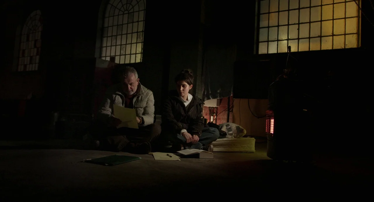 All You Need Is Death (2023) movie still: wide — A dimly lit basement room with concrete floors and large high-set industrial windows. Two people…