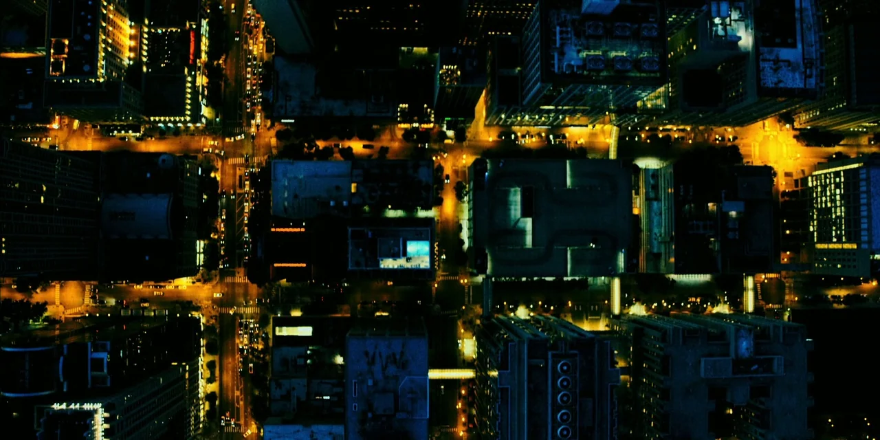 Polar (2019) movie still: establishing — A dense urban grid of high-rise buildings and intersecting streets appears at night. Numerous yellow…