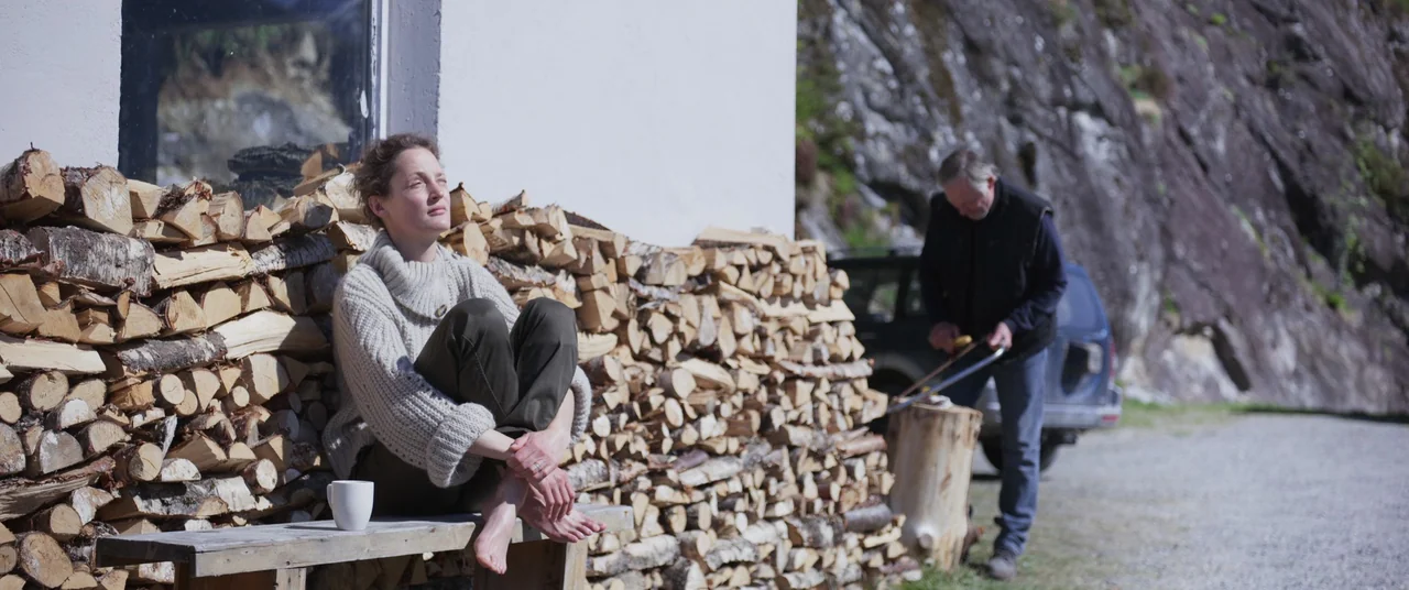 More Than Ever (2022) movie still: medium — A woman rests on a bench against a tall stack of firewood. An older man stands behind her using a hand…