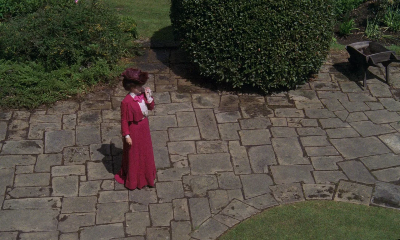 A Room With A View (1985) movie still: fullbody — A stone paved area features a large hedge and a metal wheelbarrow nearby. A woman in 1900s attire…