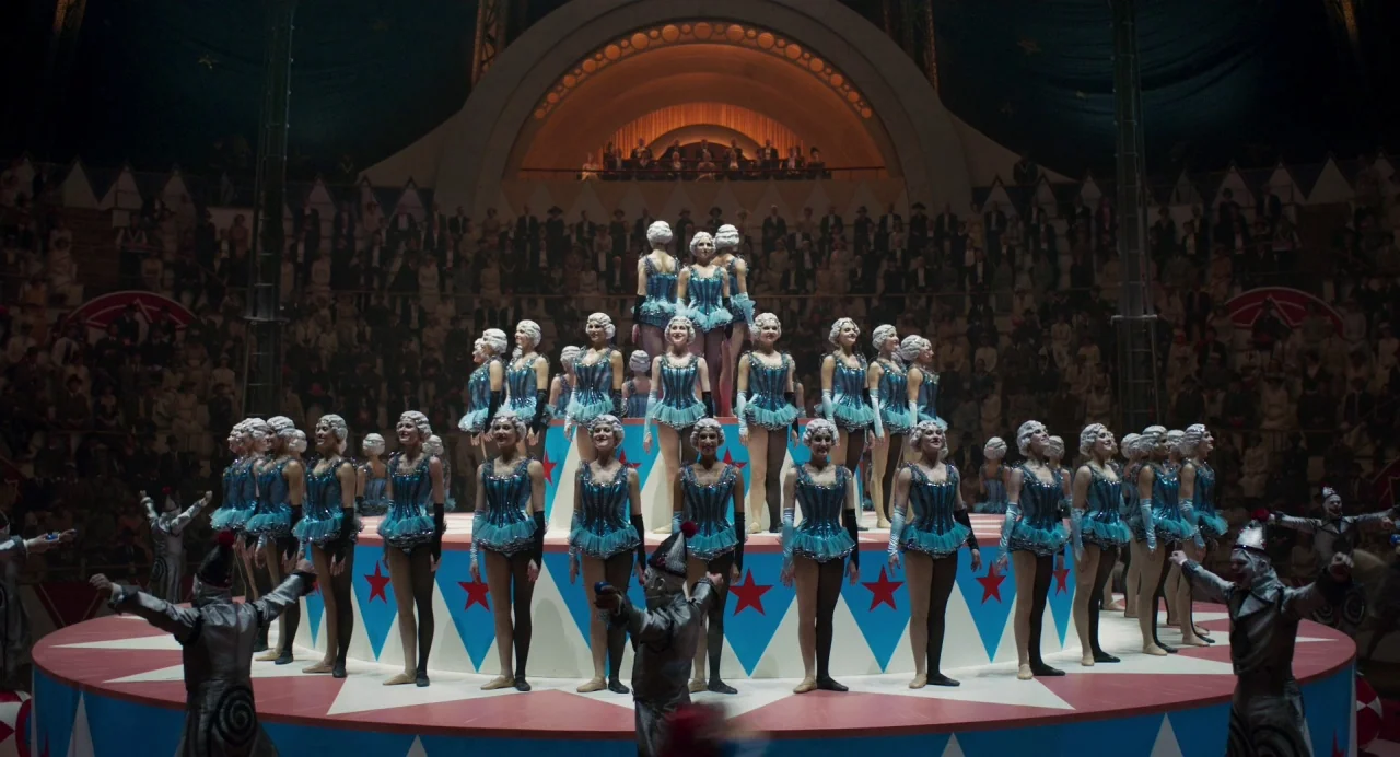 Dumbo (2019) movie still: wide — A tiered circular stage inside a large tent with a seated audience watching. Performers in blue costumes stand in…